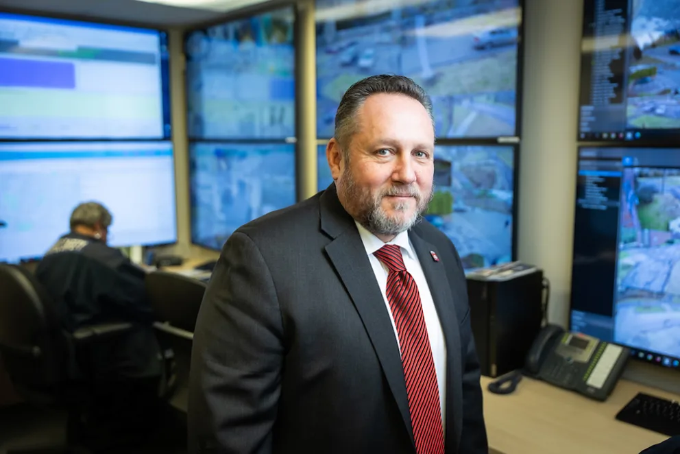 A person in a suit with a red tie stands in a control room with multiple monitors displaying surveillance footage. Another person works at a desk.