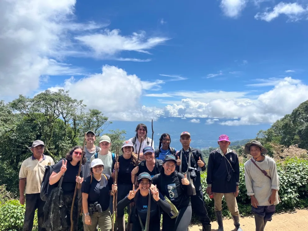 Group of people with walking sticks pose on a scenic hilltop with trees and clouds in the background.