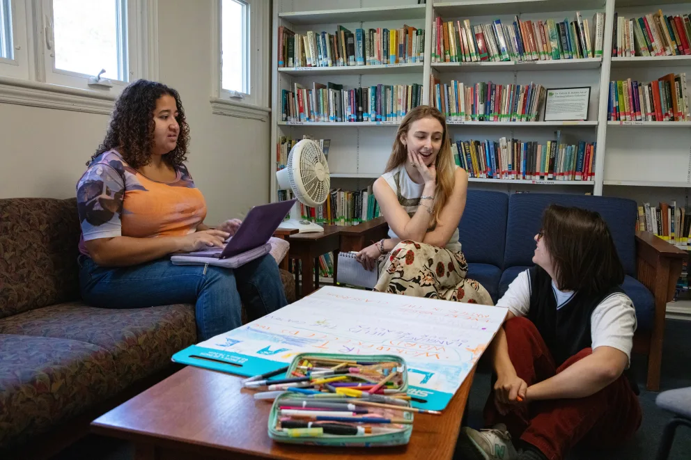 Three people in a library lounge area, one using a laptop, another sitting on the floor, and one smiling, with a large paper and markers on a table.