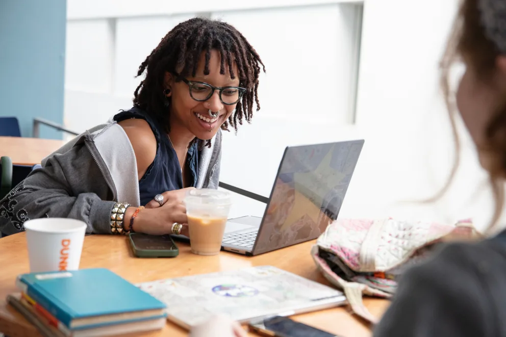 Person with dreadlocks and glasses smiling at laptop in a cafe. Coffee and books on table, colorful backpack nearby.
