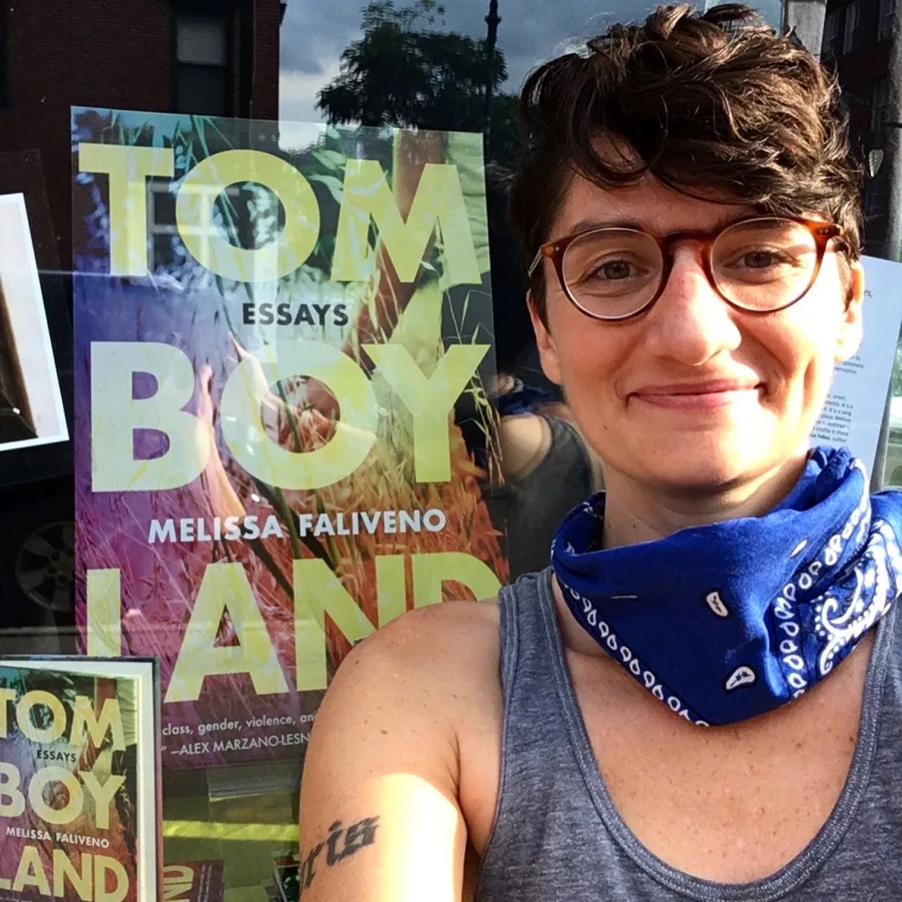 Person with glasses and a blue bandana smiles in front of a "Tomboyland" poster by Melissa Faliveno.