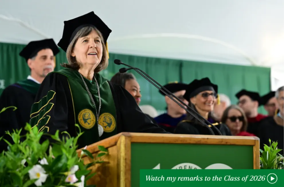 Person in academic regalia speaks at a podium during a graduation ceremony, with others in similar attire in the background.