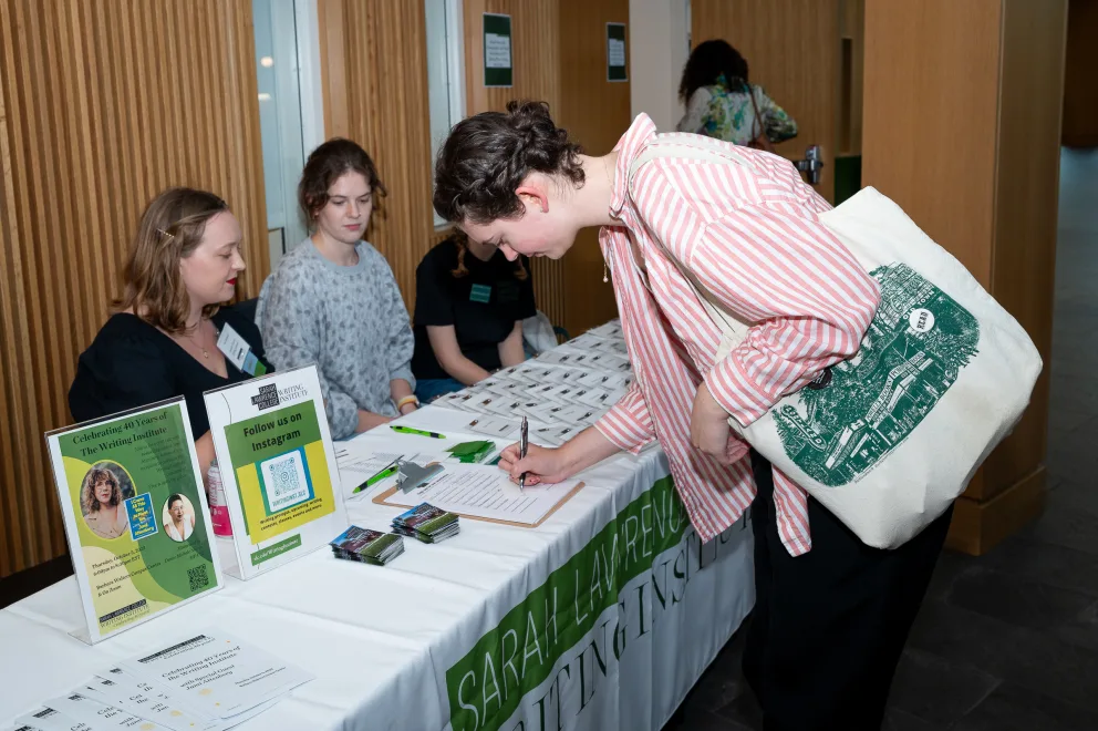 a woman signing a book at a table with people sitting at it and a tote bag in front of her