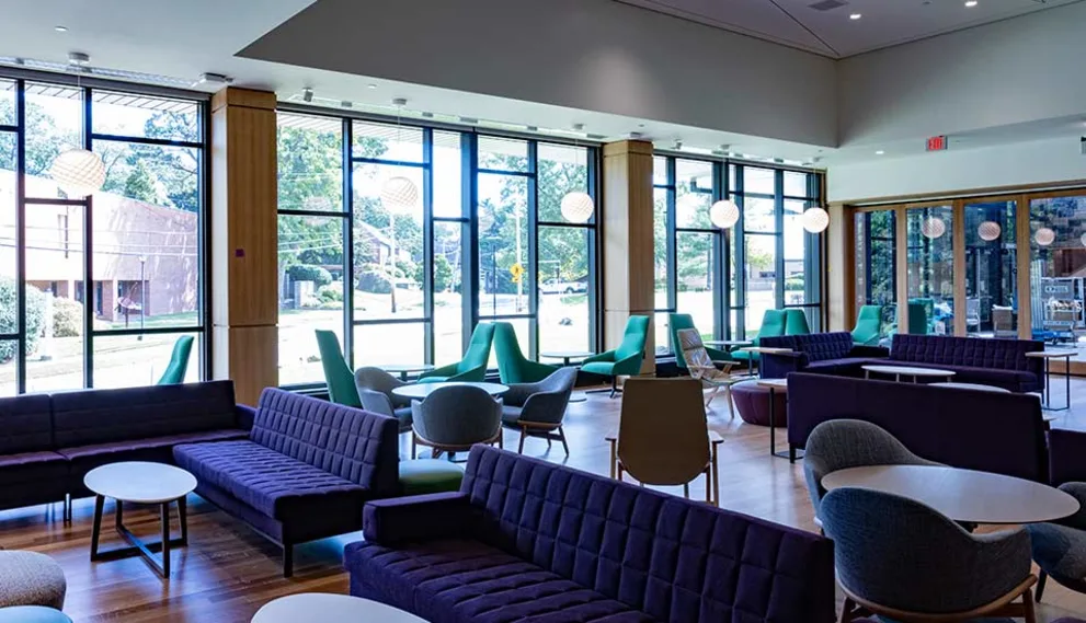 Modern lounge area with dark sofas, round tables, and large windows showing a view of trees and a building outside.