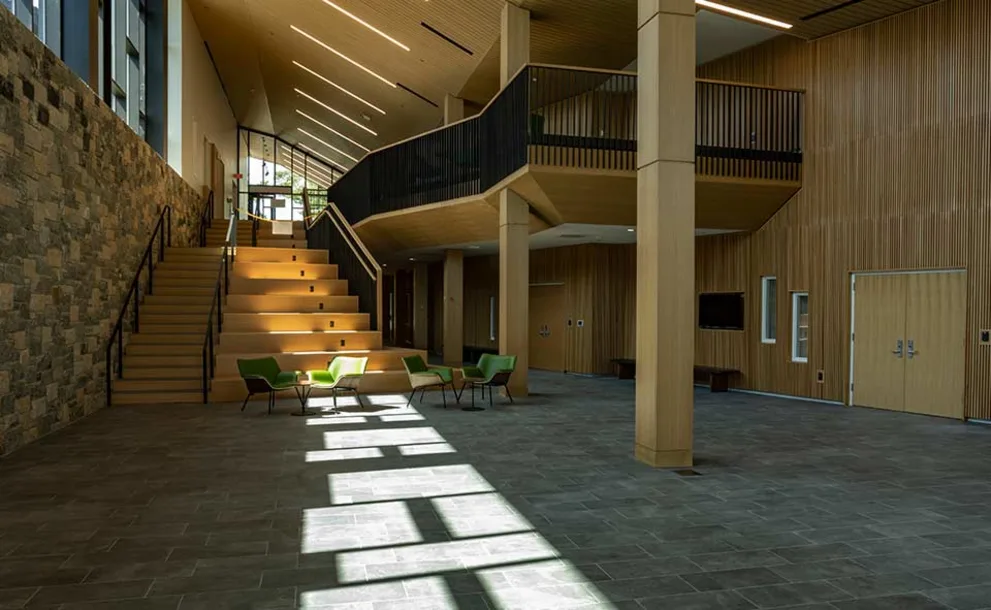 Spacious modern lobby with high ceilings, large windows, stone wall, and a wooden staircase. Green chairs and tables are arranged on the right.