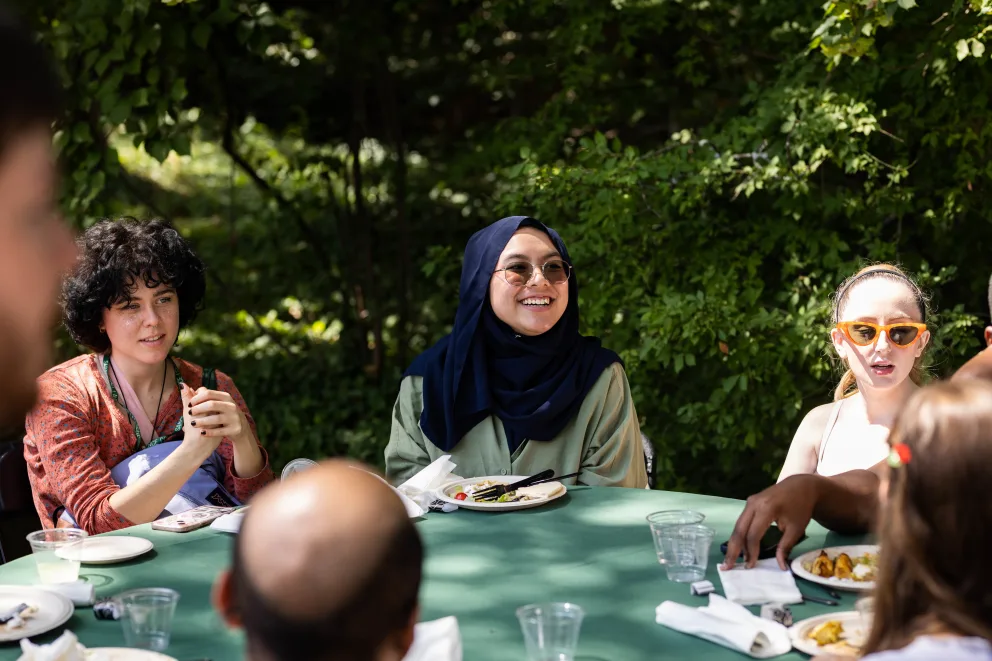 Group of people seated at a table outdoors, smiling and talking, surrounded by trees. Plates and glasses are on the table.