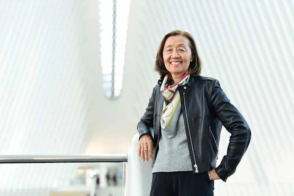 Person smiling, wearing a leather jacket and scarf, stands in a modern, bright interior with white, vertical lines in the background.