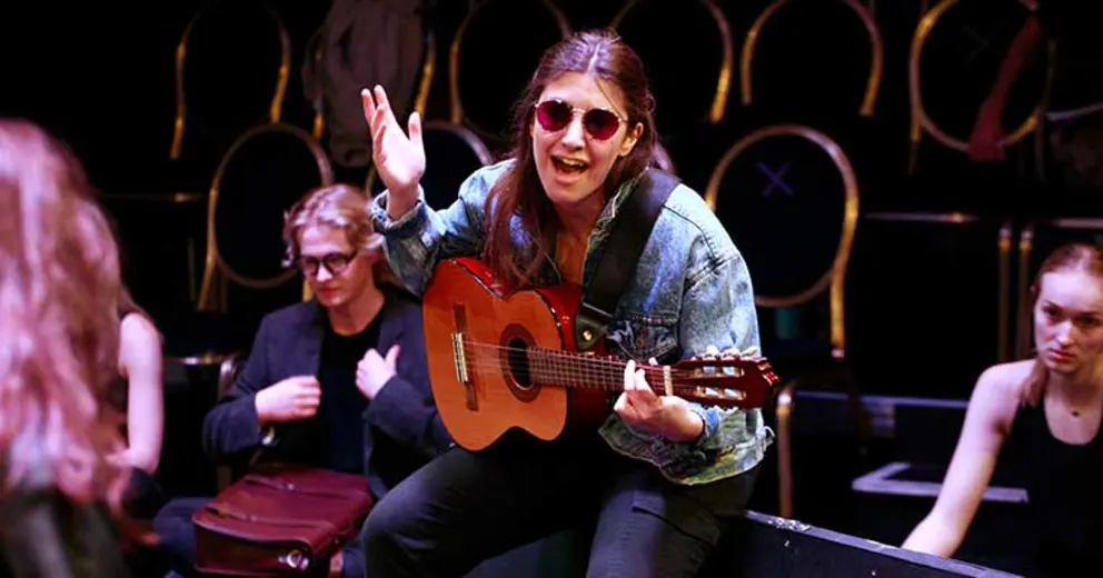Person wearing sunglasses plays a guitar and sings passionately. Others sit nearby with chairs in the background.