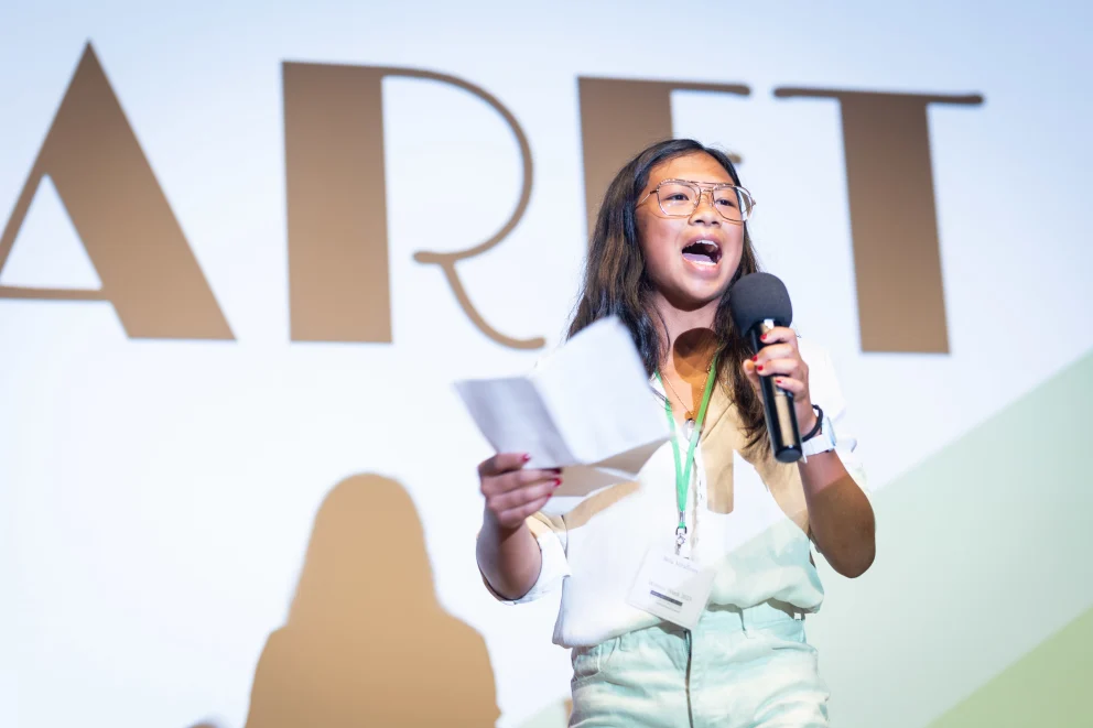 Person speaking into a microphone on stage, holding papers, with bold letters in the background.
