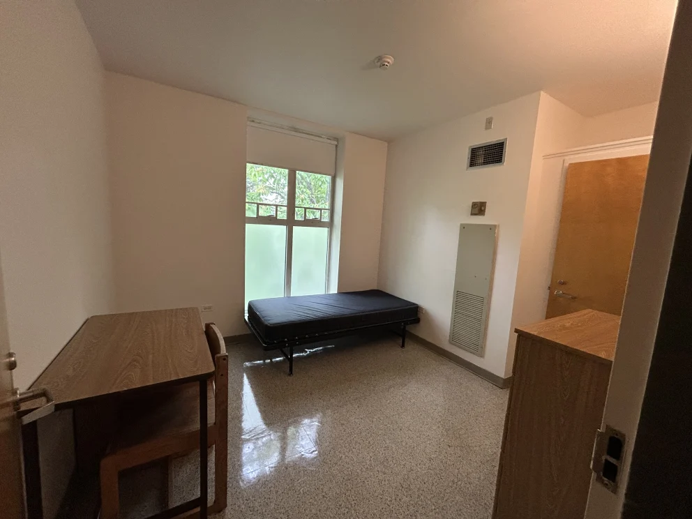 Empty dorm room with a metal-framed bed, wooden desk, and chair. A window with frosted glass and a closed wooden door are visible.