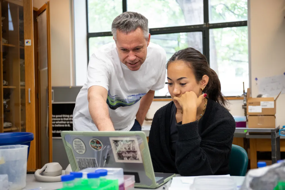 Person pointing at a laptop screen, guiding another person seated and focused on the screen in a lab or classroom setting.