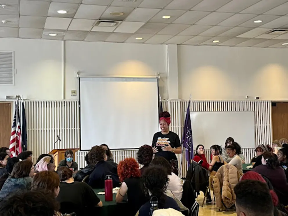 A man is standing in front of a large screen, speaking to a group of people in a room.