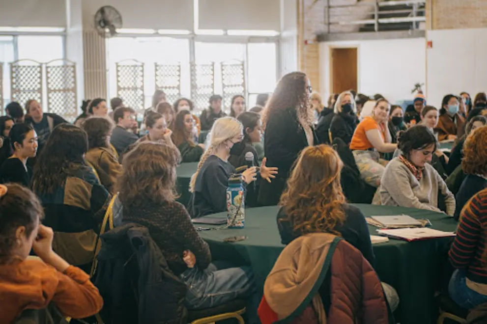 A large group of people is gathered in a room, sitting at tables and chairs, with some standing around. They are engaged in conversations and enjoying the event. There are multiple chairs and tables, with some people sitting on them, and a few people standing. The atmosphere is lively and social.