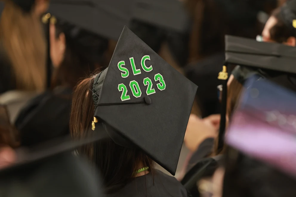 Graduation cap with "SLC 2023" in green letters, surrounded by others wearing caps and gowns at a ceremony.