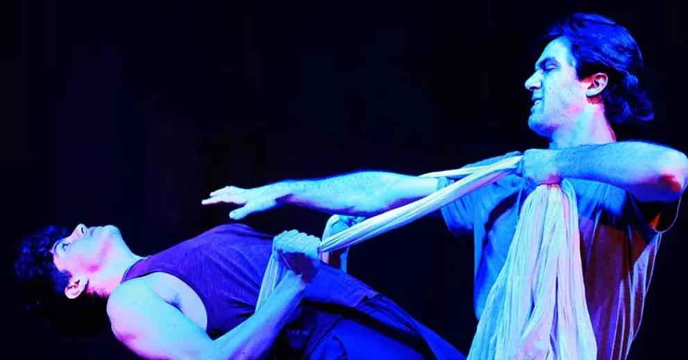 Two people engage in a dramatic performance under blue lighting, using fabric as part of their interaction.