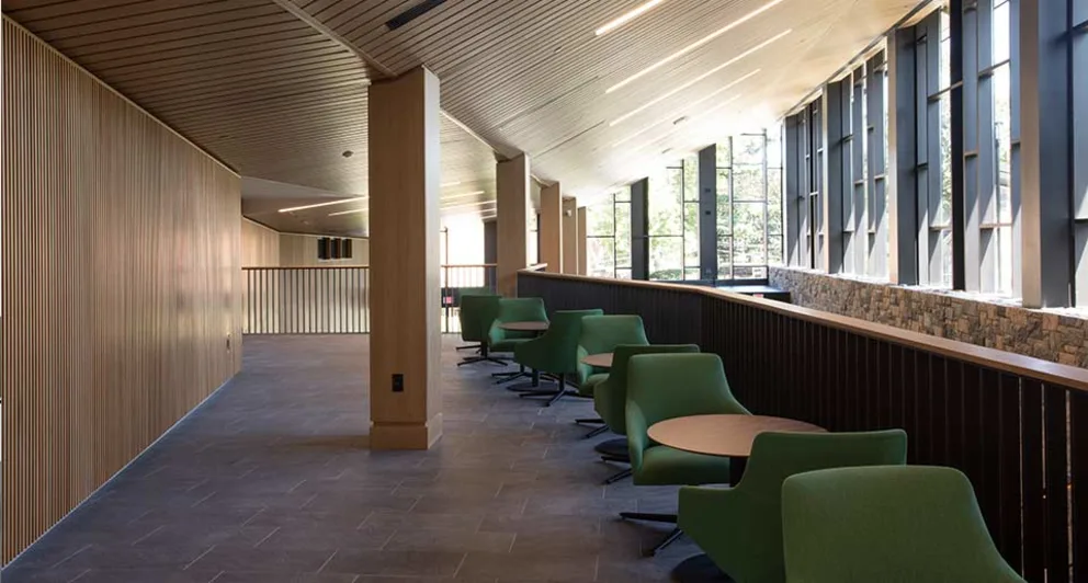 Modern lounge area with green chairs, round tables, wood paneling, and large windows letting in natural light.
