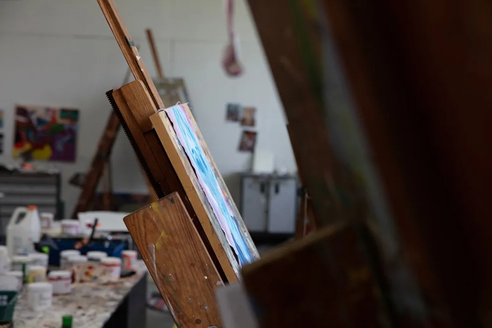 An easel with a colorful painting in an art studio, surrounded by tables with paints and supplies.