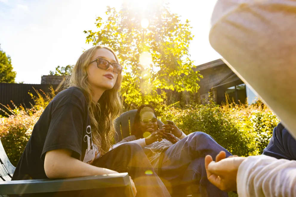 People relaxing outdoors in sunlight, surrounded by greenery. One person smiles in sunglasses, while another looks at a phone.
