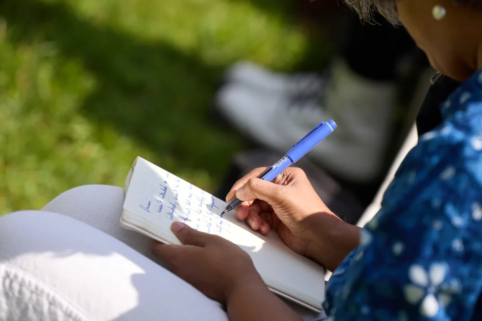 Person writing notes with a blue pen in a notebook on their lap, sitting outdoors on grass.