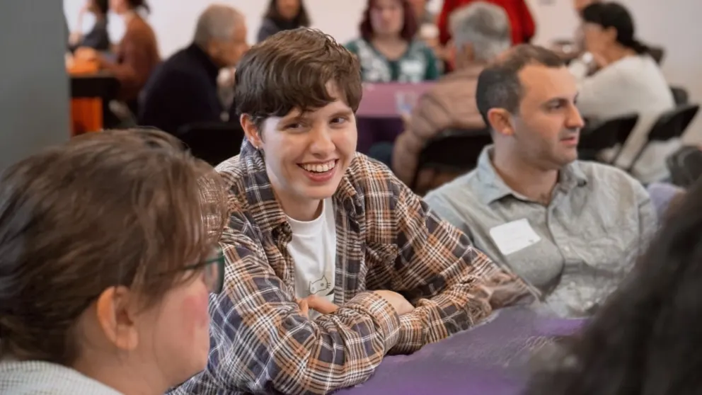 Person in a plaid shirt smiling at a table with others in a lively gathering or event.