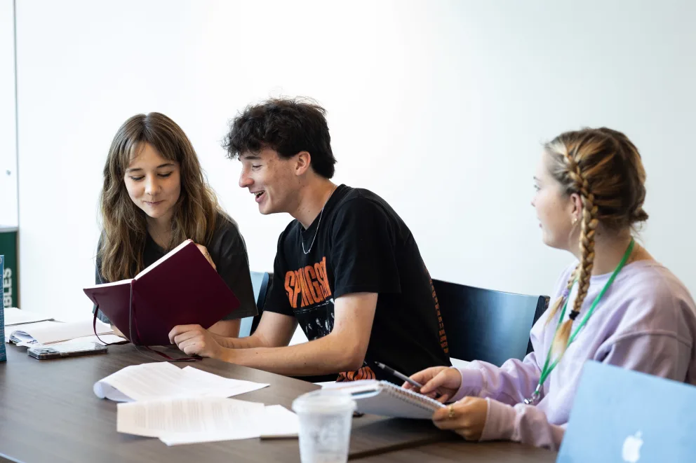 Three people sitting at a table, talking and reading notebooks. There's a laptop and papers on the table in a bright room.