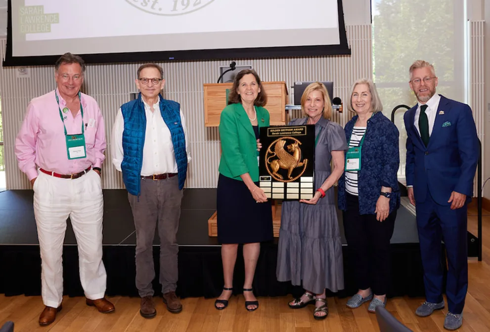 Six individuals stand on a stage, with two holding an award plaque with a decorative design. A podium and screen are visible in the background.