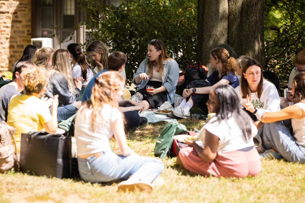 A group of people sitting on grass under a tree, engaged in conversation and enjoying a picnic.