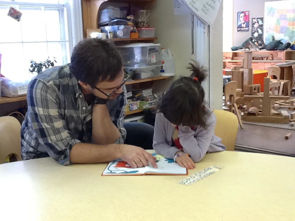 Two people sitting at a table in a classroom, reading a book together. Shelves with supplies and a bright window are in the background.