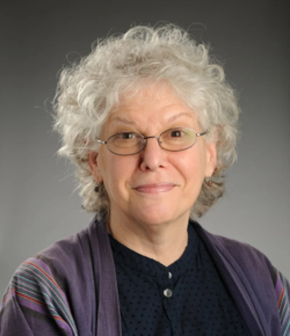 Person with curly gray hair and glasses, wearing a dark shirt and purple shawl, set against a gray background.