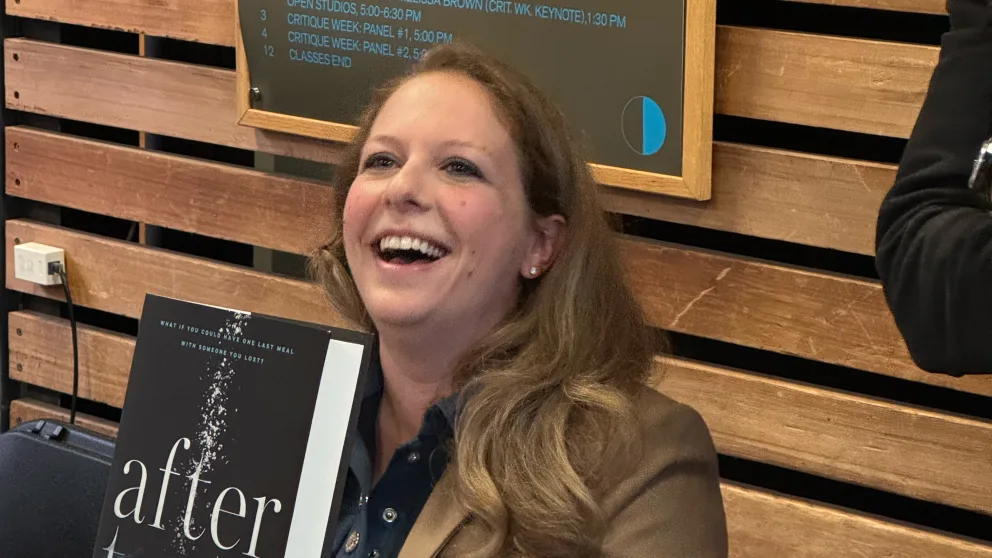 Person smiling while holding a book titled "After." They're in front of wooden slats and a schedule board.