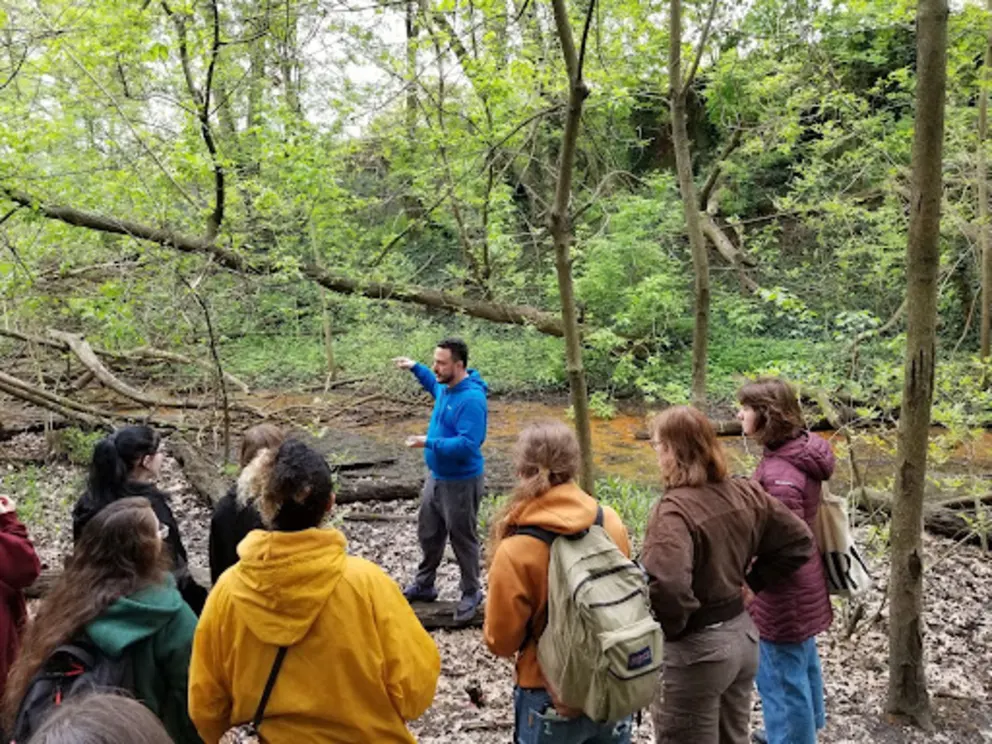 a group of people standing in a forest looking at a stream with a man in a blue jacket and a woman in a yellow jacket