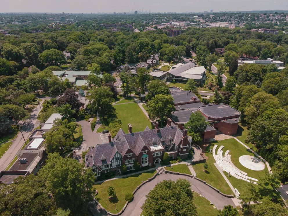Aerial view of a campus with historic buildings, green lawns, and surrounding trees, with a city skyline visible in the background.
