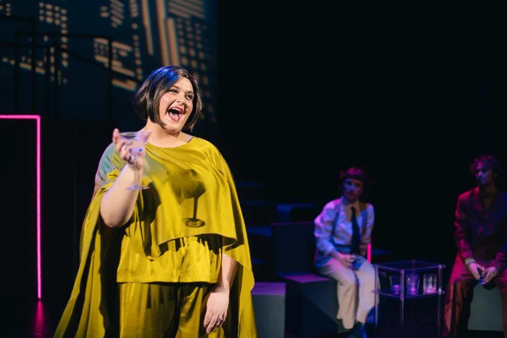 Person in a bright yellow outfit holds a glass and smiles on a stage. Two others sit in the background with a cityscape backdrop.