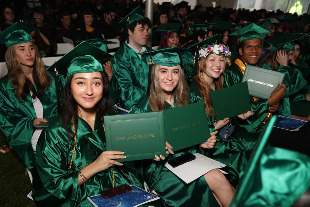 Graduates in green caps and gowns hold diplomas while seated in a crowded auditorium, smiling and celebrating their achievement.