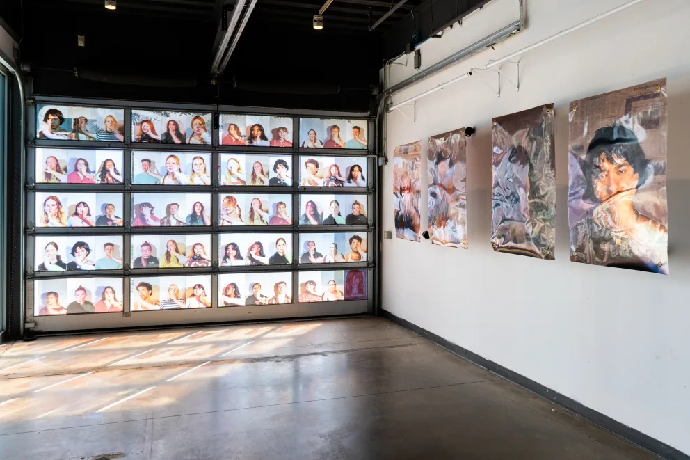 Art gallery with a wall of portraits in three rows and three large paintings on the adjacent white wall. Natural light fills the space.