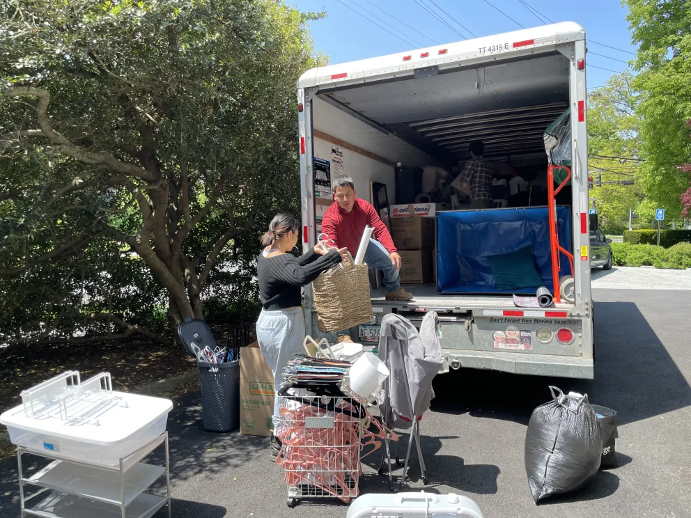 People loading various items into a moving truck on a sunny day. The truck is parked on a driveway near trees, with bags and boxes visible.