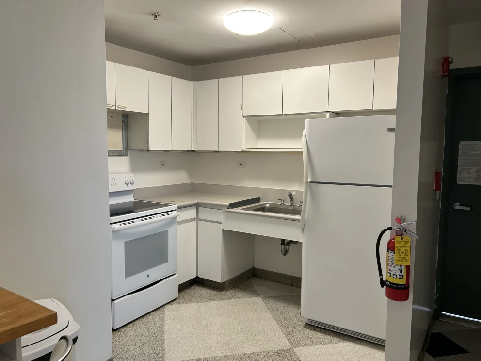 Small kitchen with white cabinets, stove, sink, and refrigerator. Fire extinguisher mounted near the door. Overhead light is on.