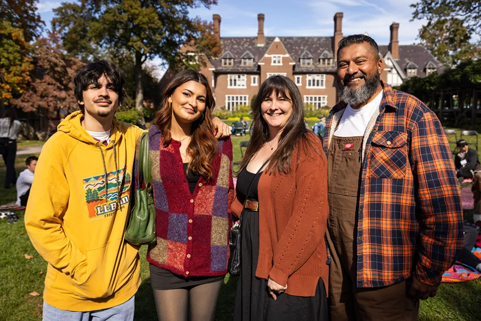 Four people smiling, standing together on a sunny day in a park with trees and a large, old-style building in the background.