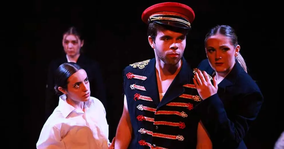 Performers in theatrical costumes on stage, one wearing a decorated jacket and red hat, flanked by two others in dark attire, against a dark background.