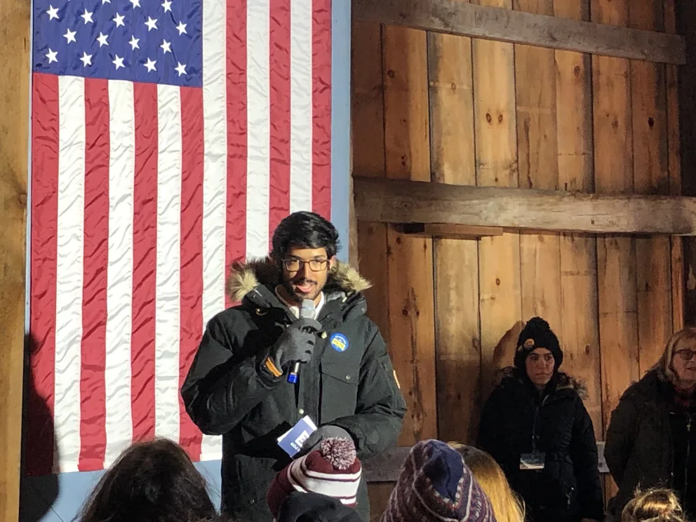 Person speaking with a microphone in front of a U.S. flag, wearing a winter coat and gloves, surrounded by onlookers in a wooden room.