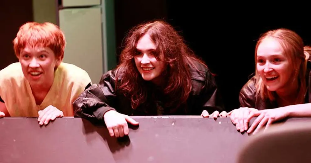 Three people leaning over a barrier, smiling and looking out enthusiastically, lit with dramatic stage lighting.