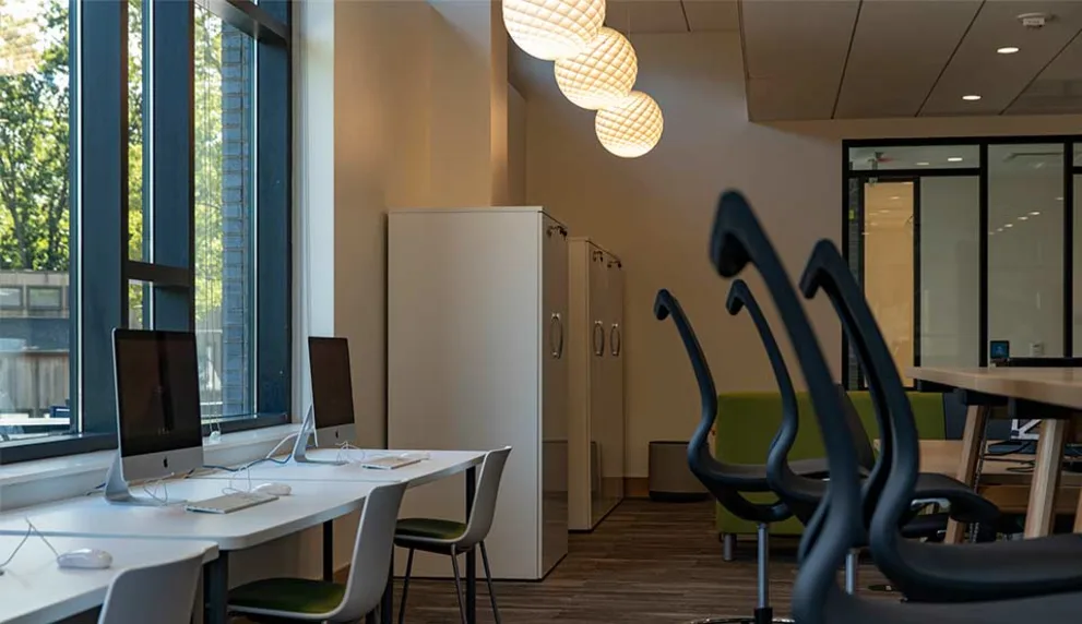 A modern office space with rows of computers on white desks, green chairs, dark office chairs, and hanging spherical lights near large windows.