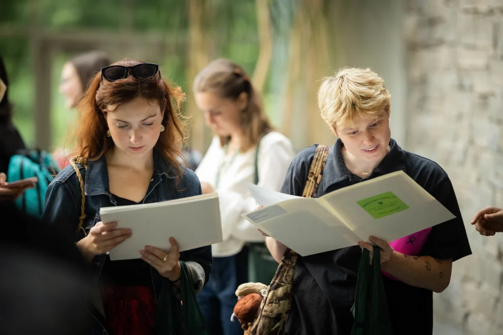 Two people reading documents in a lively, outdoor setting with others in the background.