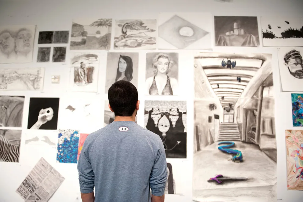 Person in a blue shirt stands in front of an art wall filled with various black-and-white and color sketches and paintings.