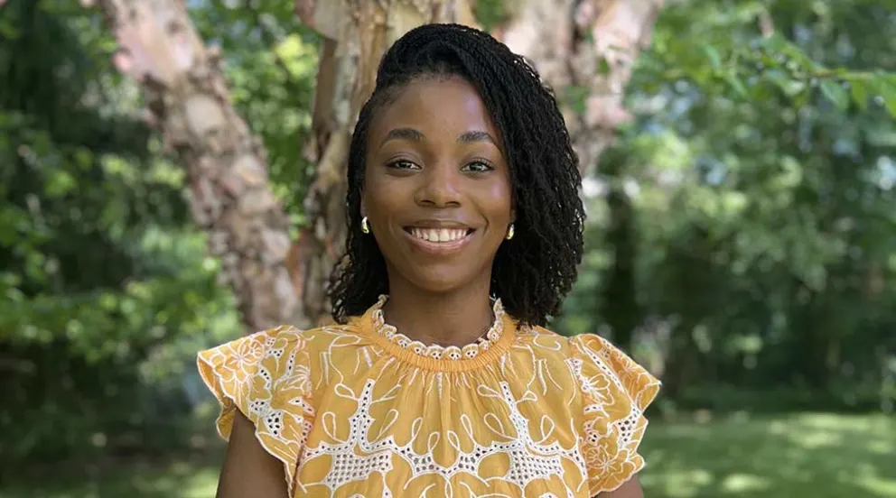 Person smiling outdoors, wearing a yellow patterned top. Trees and greenery are visible in the background.