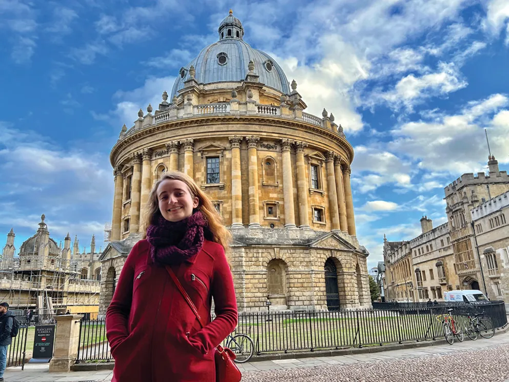 Person in a red coat stands in front of the Radcliffe Camera, Oxford, under a blue sky with scattered clouds.