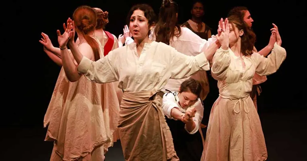 Performers on stage dressed in loose, earth-toned costumes, with arms raised, immersed in an expressive scene under stage lighting.
