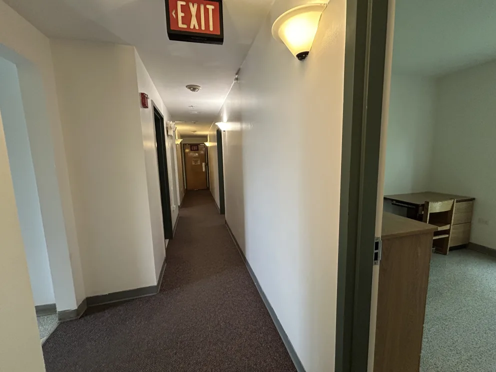 A hallway with beige walls, carpeted floor, wall lights, and an "EXIT" sign. Doorway on right leads to a room with a desk and chair.