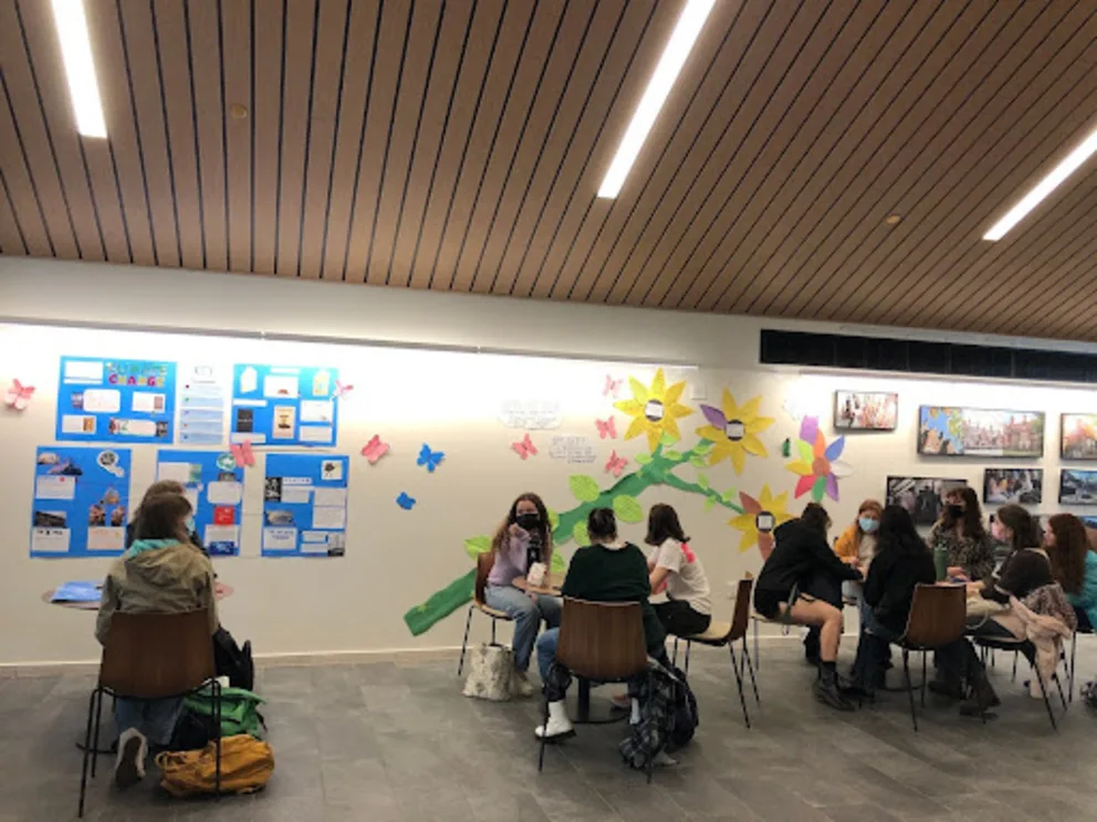 A group of people are sitting at tables in a room, with some of them looking at a large poster on the wall.