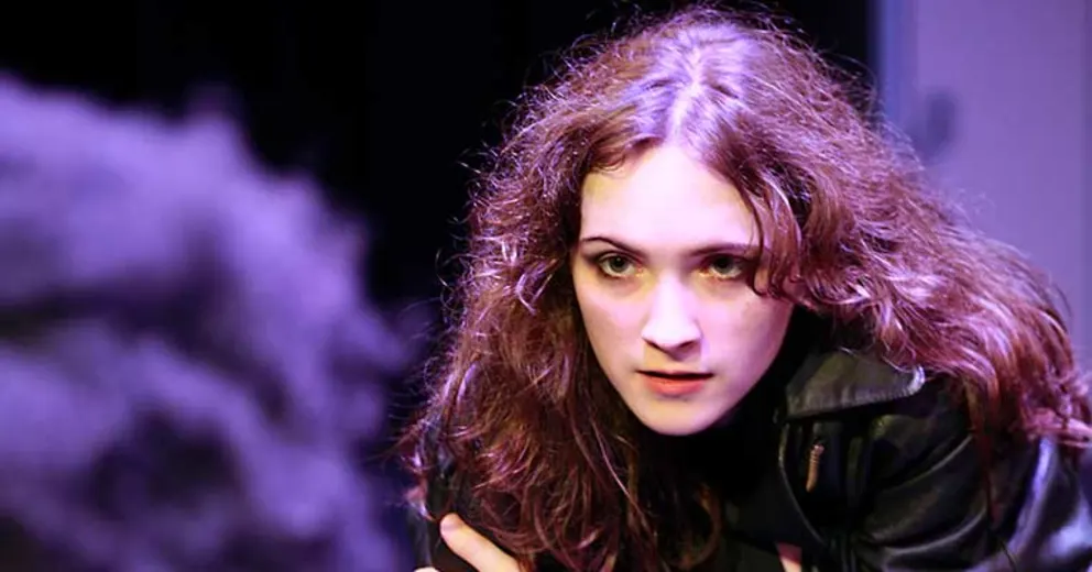 Person with long, curly hair leans forward attentively, wearing a black jacket. Background is dimly lit.